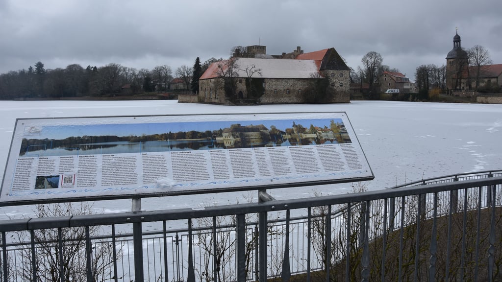 Blick über den winterlich gefrorenen Schloss-See auf das Flechtinger Wasserschloss. Im Vordergrund informiert ein Panoramaschild über markante Punkte rund um See und Ort.